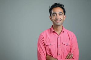 Smiling South Asian man wearing a pink button-up shirt, arms crossed, against a neutral gray background. photo