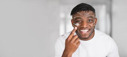 African American man smiles brightly while applying rejuvenating skin care products on his face. He enjoys the calming experience of a facial massage in a serene setting, copy space photo