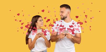 A couple shares a joyful moment on Valentine's Day, smiling at each other while holding smartphones. Colorful hearts surround them, adding to the affectionate atmosphere of their celebration. photo