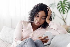 Upset african american girl using smartphone while sitting on couch at home, having depression, waiting for importrant call, free space photo