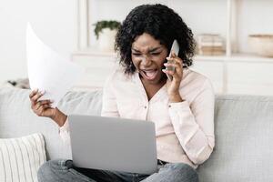 Furious black woman talking on cellphone while working with papers and laptop at home, having business problems, arguing with somebody by phone photo