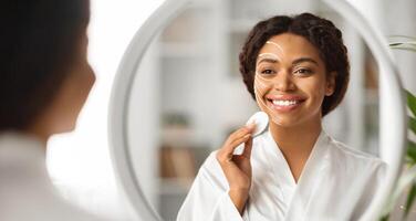 African American woman is engaged in her skincare routine, applying rejuvenating products with a smile. She looks relaxed while observing her reflection in a round mirror photo