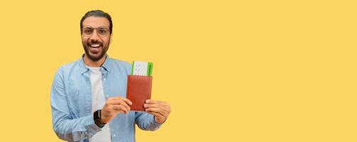 Smiling indian man presenting a passport with boarding passes on a yellow background, suggesting travel readiness photo