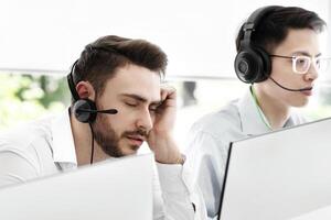 Tired call centre representative and his colleagues at work in open space office. Exhausted hotline operator sleeping at workplace, feeling fatigue or stress after communication with clients photo