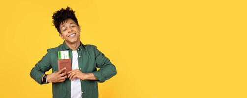 A satisfied african american guy gently holds a passport, evoking feelings of contentment and longing for travel against a yellow background photo