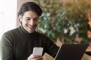 Wealthy young businessman using phone while working with laptop at cafe, time for entertainment, copy space photo