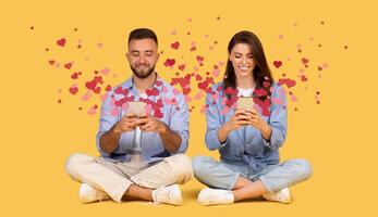 A joyful couple sits together on a yellow background, each holding a smartphone while smiling. They express affection and connection through online messages filled with hearts, celebrating St. photo