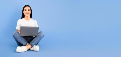 Technology. Portrait of a smiling young woman sitting with legs crossed, using laptop computer isolated over blue studio background, looking at camera. Satisfied lady studying or working with notebook photo