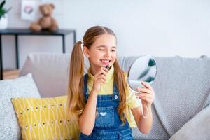 Everyday Make Up. Portrait of cheerful teenage lady applying lipstick, holding mirror and looking at herself with smile, sitting on the couch at home, getting ready for school or date photo