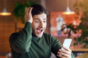 Thrilled guy got shocking news, looking at smartphone and holding head, sitting at cafe and drinking coffee, empty space photo