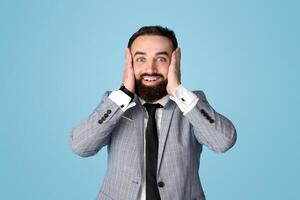 Portrait of shocked young businessman holding his head in excitement, cannot believe his luck or success on blue studio background. Positive millennial CEO feeling astonished and amazed photo