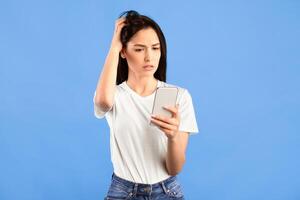 Dissatisfied gloomy female model looking at smartphone while browsing social media or texting sms and being disappointed, standing over blue background. Woman trying to call but there is no signal photo