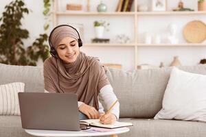Young arabic woman in hijab watching webinar on laptop and taking notes while sitting on couch at home, free space photo
