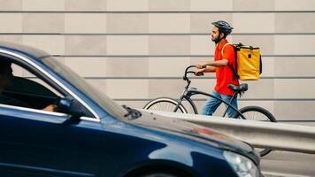 Delivery by courier at transport in city. Guy with beard in helmet and with backpack, going through city with bicycle on gray brick wall background, side view, free space photo
