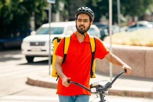 Delivery to home or office. Young man with beard in protective helmet with large yellow backpack with bicycle looking for the address of client, outdoors, free space photo