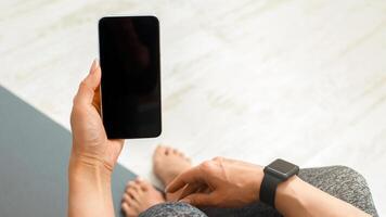Modern workout at home with gadgets. Woman in sweatpants with smartwatch, holds in hand phone with blank screen, sitting on mat on floor, pov, close up, copy space photo