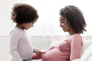 Curious little black girl touching her pregnant mother's belly, sitting on couch at home together, side view with free space photo