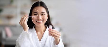 Asian woman is happily applying a serum to her face using a dropper while sitting in a stylish, well-lit room. She enjoys her skincare routine with a focus on rejuvenation and pampering, copy space photo