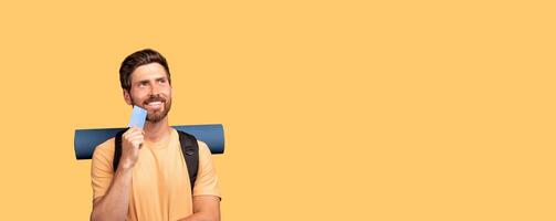 A millennial man traveler holds a blue card, reflecting on trip possibilities. He wears a casual t-shirt, has a backpack, and is set against a bright, isolated background. photo