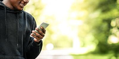 Positive black man with wireless headset using mobile phone, coming back from training at park, empty space photo