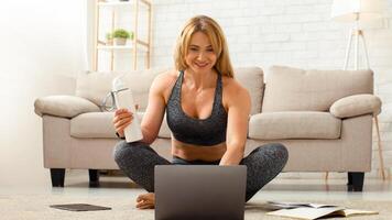 Coach works from home. Smiling woman holds bottle and looks at laptop, sitting on floor, free space photo