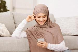 Nice App. Happy Muslim Girl In Hijab Using Smartphone At Home, Checking New Application, Browsing Internet Or Messaging With Friends, Closeup photo