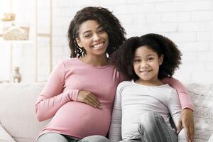 Happy pregnant african woman and her little daughter relaxing on couch at home and smiling to camera, hugging and smiling, copy space photo