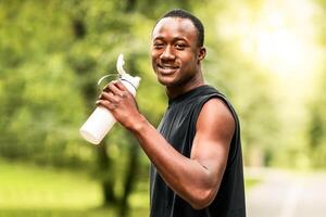 Positive african runner having break while exercising at park, drinking water, copy space photo