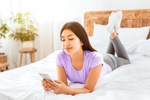 Chinese Woman With Smartphone Using Mobile Application Or Texting Lying On Bed At Home. Modern Communication photo