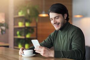 Young cheerful entrepreneur using mobile phone while drinking coffee at cafe, empty space photo