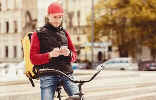 Delivery Guy On Bicycle Using Phone Waiting For Food Order In Delivering Mobile Application Standing Outside In City. Empty Space photo