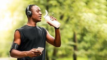 African jogger drinking water and using smartphone, resting while training at park, copy space photo