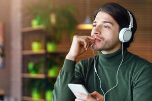 Pensive young man in headphones listening to music, using mobile phone, drinking coffee, cafe interior, empty space photo