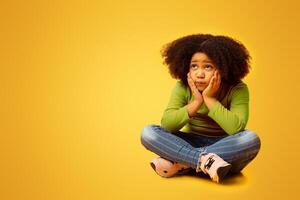 Bored cute little afro girl sitting on floor, resting head on hands and thinking about something, looking away at copy space over yellow background photo