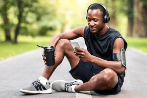 Morning exercising. Happy athletic black guy resting on path at public park, using phone and drinking water, copy space photo