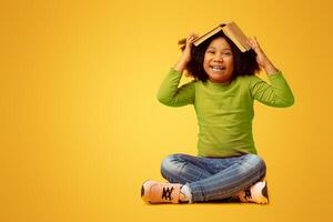 Education Concept. Portrait Of Playful Little Afro Girl With Opened Book On Head Sitting On Yellow Background In Studio And Laughing, Copy Space photo