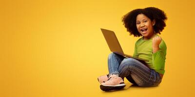 Joyful little african american girl sitting with laptop on lap and celebrating success, raising fist and emotionally shouting on yellow background photo