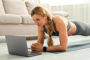 Sport online and watching . Middle aged woman in sportswear with fitness tracker makes plank at mat on floor and looks at laptop in interior of living room, copy space photo