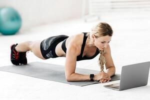 Online lesson at home during coronavirus pandemic. Attractive mature sportswoman in sports uniform makes plank on mat with fitness ball and bottle of water on floor and looks in laptop, free space photo