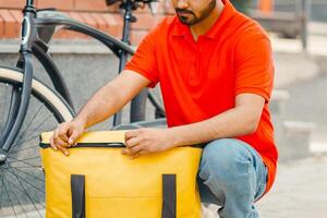 Accurate delivery. Busy bearded courier zips up large yellow bag for client, beside a bicycle on street, outside, free space photo