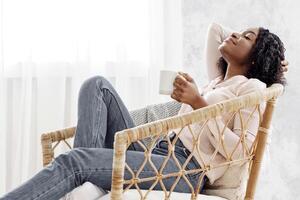 Home Harmony, Young African Lady Relaxing With Cup Of Coffee In Modern Wicker Chair, Drinking Tea, Sitting With Closed Eyes Near Window, Enjoying Lazy Weekend Pastime, Side View With Copy Space photo
