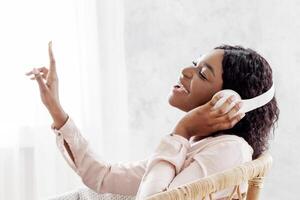 Joyful African Woman Singing While Listening Music With Wireless Headphones At Home, Sitting In Stylish Wicker Chair, Enjoying Favorite Song Playlist, Dancing With Hand, Side View With Copy Space photo