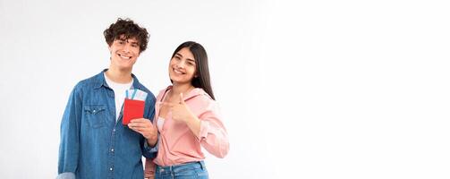 Cheerful young couple posing with tickets on white studio background, girlfriend pointing at guy's passport and boarding pass traveling together, smiling to camera. Travel offer advertisement photo