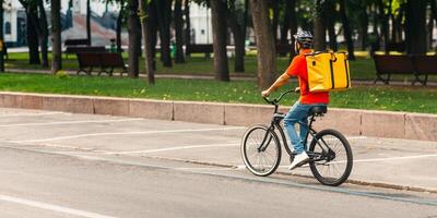 Courier bicycle delivery service. Man in safety helmet with large yellow backpack ride along road in park at city, free space photo