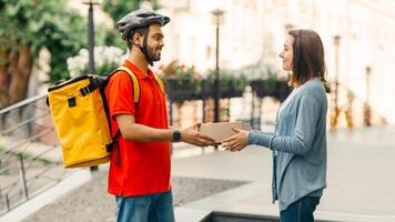 Woman takes online purchase from courier. Deliveryman in helmet, with yellow backpack gives box to client on street, near house photo
