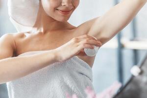 Beautiful young woman applying deodorant on armpit in front of mirror at bathroom photo