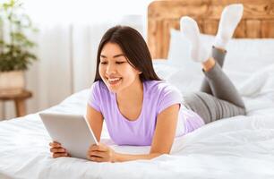 Korean Lady Using Digital Tablet Reading Books Online Lying On Bed At Home Relaxing On Weekend. photo
