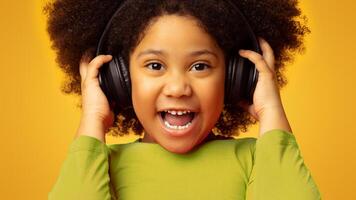 Closeup Portrait Of Excited Little African Girl Enjoying Listening Music In Wireless Headphones, Posing Over Yellow Background And Looking At Camera photo