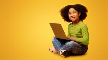 Online Education Concept. Adorable Little Afro Girl Sitting On Floor With Laptop And Looking At Camera, Posing Over Yellow Background With Empty Space photo