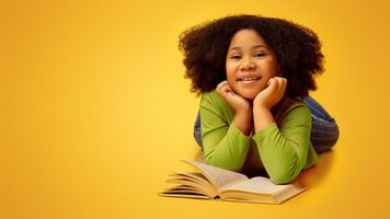 Pretty Little Afro Girl Lying On Floor With Book, Reading And Enjoying To Study, Yellow Background With Free Space photo